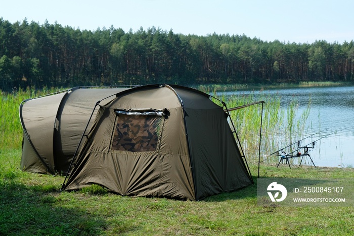 The anglers’ tents stand by the lake. There are two fishing rods on the stands above the water.