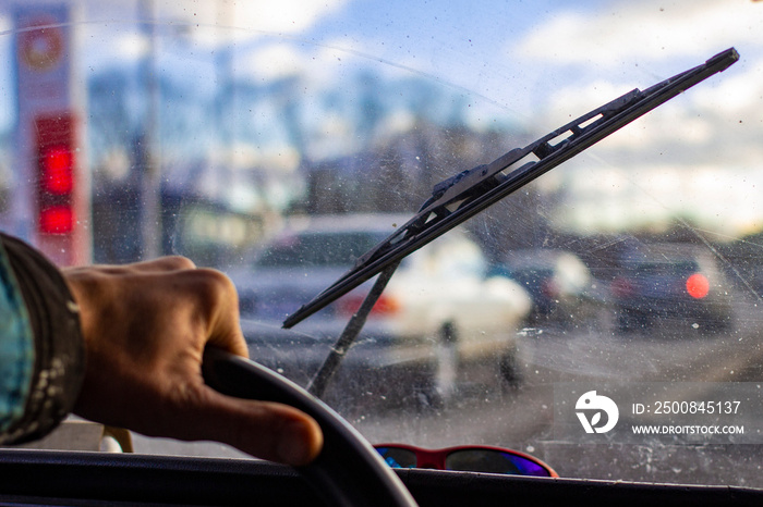 dirty scratched car windshield with wiper through blurred steering wheel with driver’s hand on blurred background