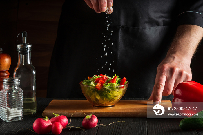 Chef or cook sprinkles salt salad of fresh vegetables on wooden table. Preparing healthy food on the kitchen