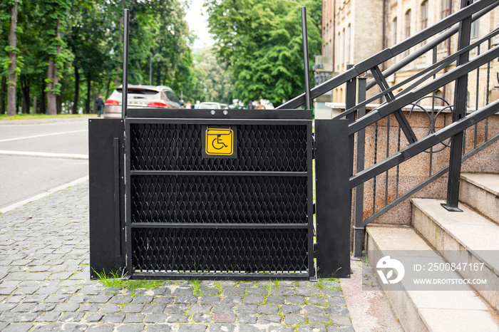 Electric platform lift at building staircase for disabled people with wheelchair sign plate on old city street. Elevator stairlift ramp mechanism for senior disability people. Medical urban equipment