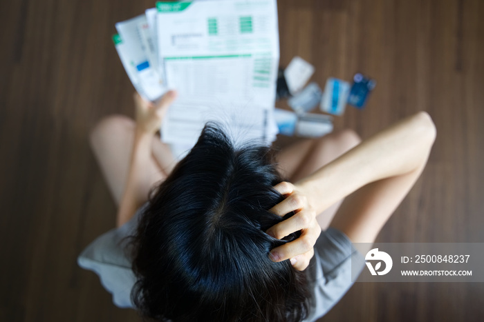 Top view of stressed young Asian woman holding bills and thinking about  finding money to pay credit card debt and all bills. She is holding head by another hand. Financial problem concept.