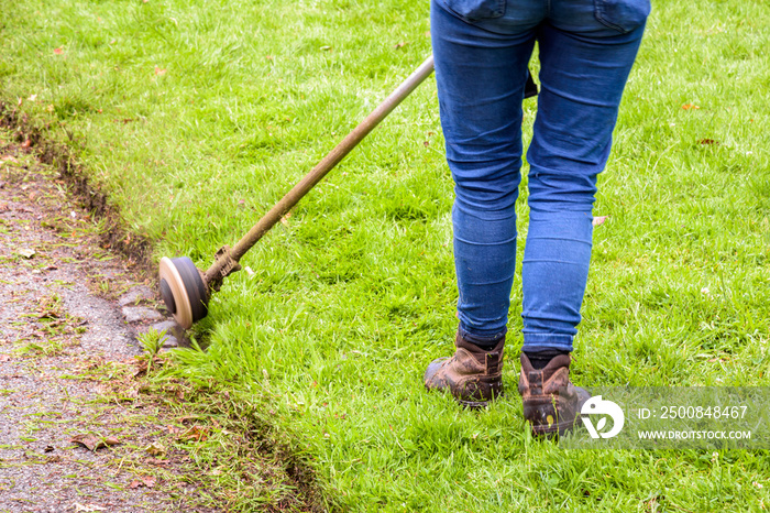 A woman is using a string trimmer to edge the lawn along the driveway.