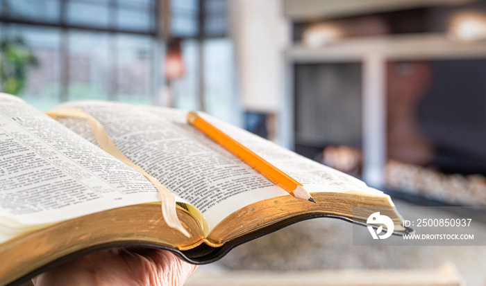 A man holds a Bible with a pencil, against the background of the living room. Reading a book in a cozy atmosphere.