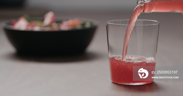 pour pink fizzy drink in a tumbler glass on walnut table with salad on background