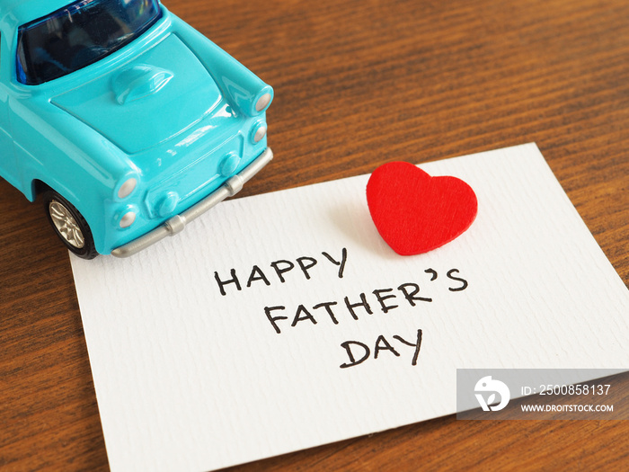 Handwriting the word  Happy Father’s Day  on white card with a red heart and blue toy car over wooden background..