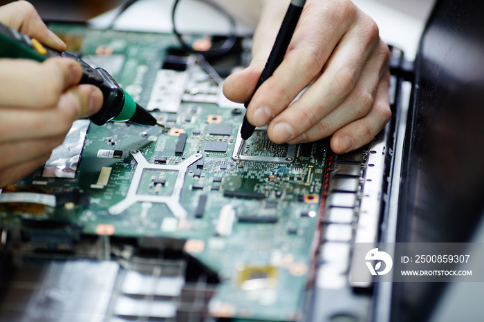 Closeup shot of male hands testing electric current in circuit board of disassembled laptop using multimeter tool on table in maintenance shop