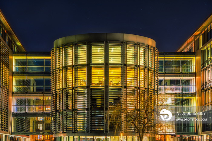 Modern opulent cylindrical office or headquarter illuminated at night with sun blinds or sunblinds and side passageways features a contemporary massive facade or imposing exterior with glazed halls