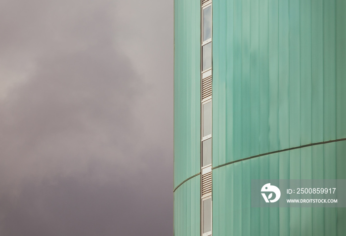 View of the green facade of a tall modern building with a vertical row of narrow windows and the cloudy sky on the left