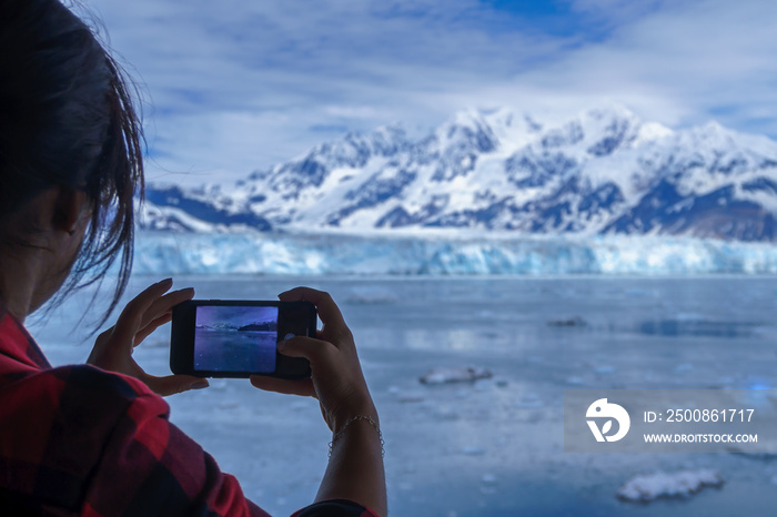 Woman is wearing a lumberjack shirt and taking a picture of Hubbard Glacier on her mobile phone. This is beautiful Alaska and you can see it on the screen of smartphone. Snowy mountains and iceberg.