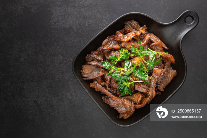 Thai food, fried sun dried beef with crispy basil leaves in black pan on the dark tone texture background with copy space for text