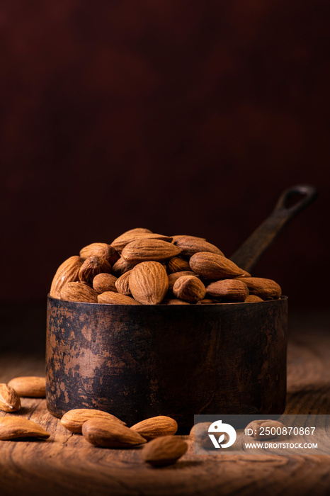 In the foreground, a group of shelled almonds in an aged metal measuring cup against a dark background