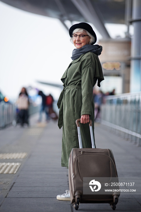 Ready to travel. Full length of positive senior woman wearing hat is standing outdoors with suitcase while looking at camera with joy. She is locating at international airport