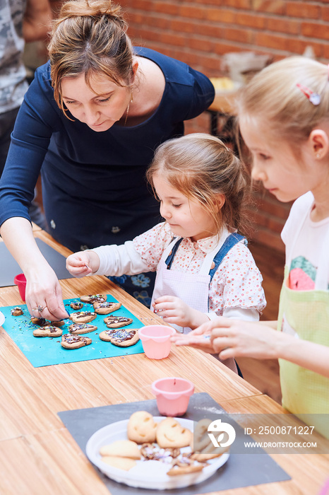 Little girl with her mom’s help decorating baked cookies with colorful sprinkle and icing sugar. Kid taking part in baking workshop. Baking classes for children, aspiring little chefs