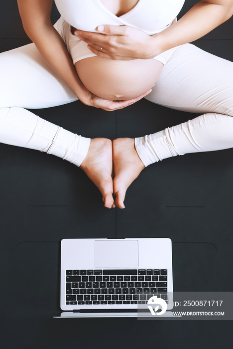 Closeup belly of pregnant woman practicing yoga online with laptop.