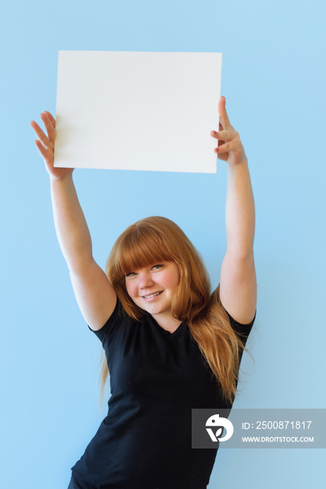 young woman of Caucasian appearance with red hair holds a poster with copyspace with two hands, she smiles, on an isolated blue background, dressed in black casual clothes