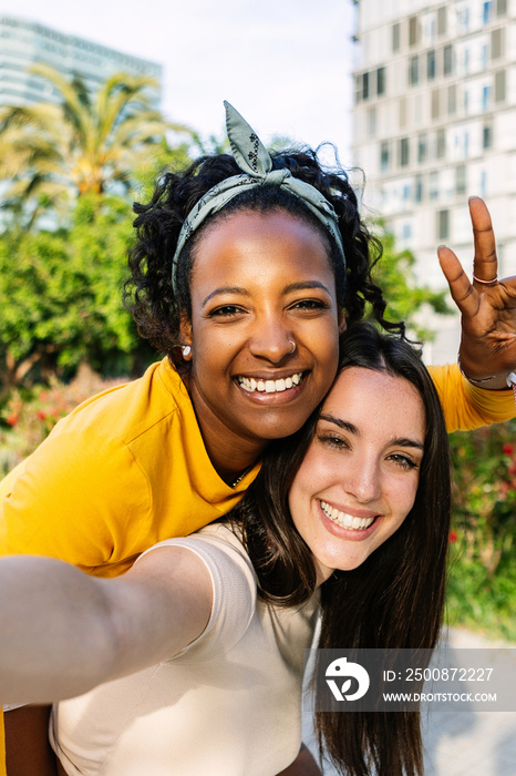 Vertical shot of two multiracial best female friends in casual summer clothes having fun together taking self portrait using phone outdoors.