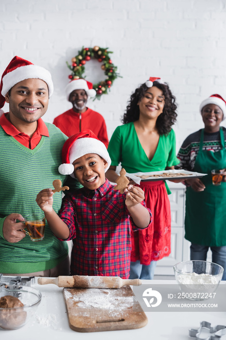 cheerful african american boy showing baked christmas cookie near family in santa hats in kitchen