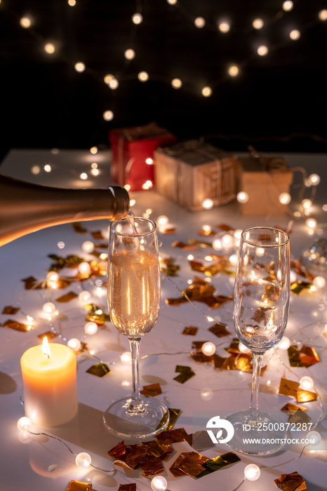 Two flutes and burning candle on table among golden confetti and lit garlands