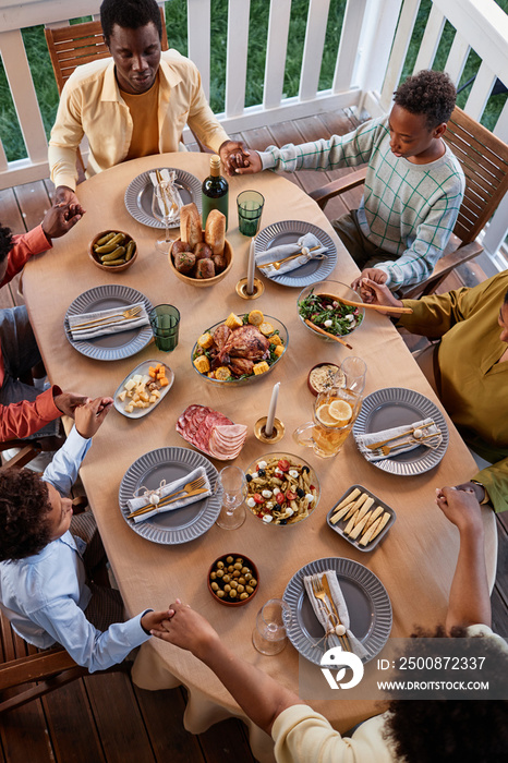 Top view of African American family saying grace at festive dinner table outdoors and holding hands in cozy setting