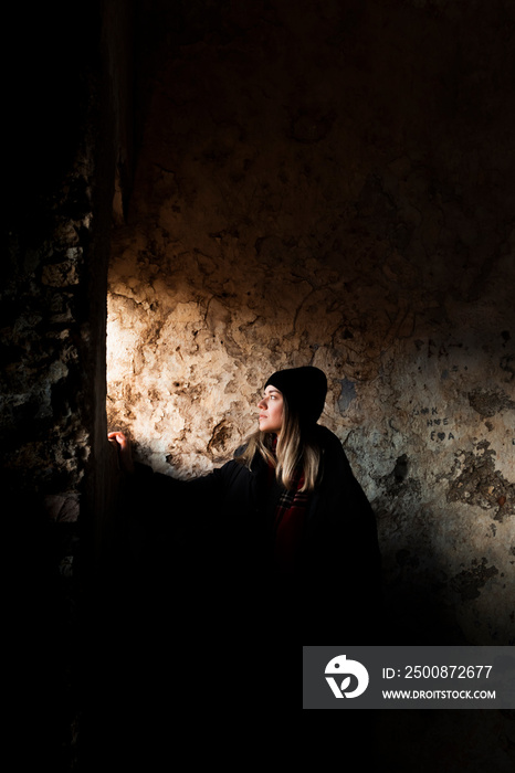 A girl in front of the window in the dark historical abandoned house. Sun lights coming from outside to girls face. A mysterious and promising atmosphere.