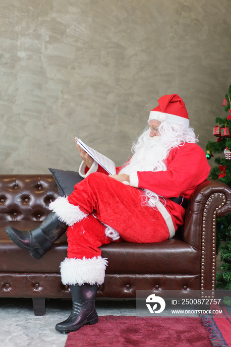 Senior man in Santa Claus costume reading newspaper on sofa in the living room at home