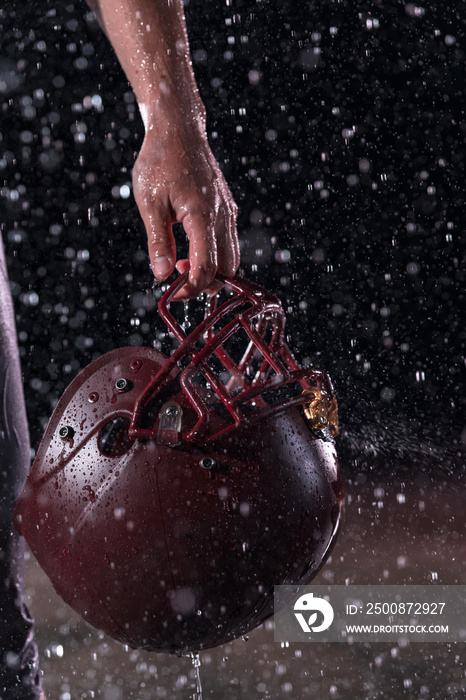 Close up of American Football Athlete Warrior Standing on a Field focus on his Helmet and Ready to Play. Player Preparing to Run, Attack and Score Touchdown. Rainy Night with Dramatic lens flare and