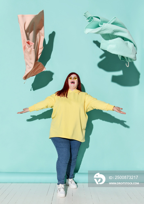 Young happy red haired overweight woman in casual clothing and sneakers playing with flying blue and pink hoodie