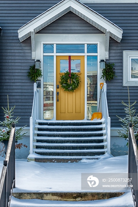 The exterior entrance to a modern blue grey colored wooden house. The yellow door has a glass window with a Christmas wreath hanging from it. There’s a snow shovel on the steps of the residence.
