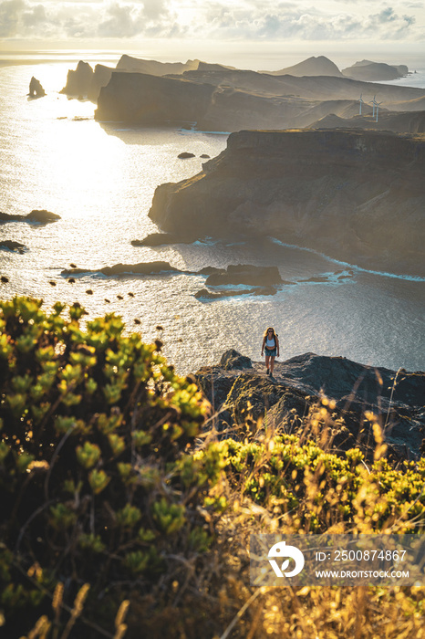 Backpacker woman enjoys enjoys hike at steep cliff over the seascape and along the rugged foothills of Madeira coast at sunrise. Ponta do Bode, Madeira Island, Portugal, Europe.