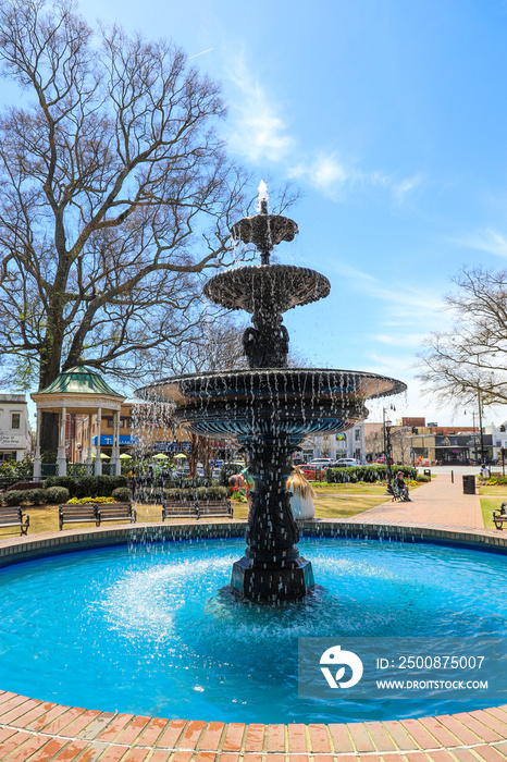 a gorgeous circular water fountain surrounded by people, black metal benches, lush green trees, buildings and bare winter trees with blue sky and clouds at the Marietta Square in Marietta Georgia USA