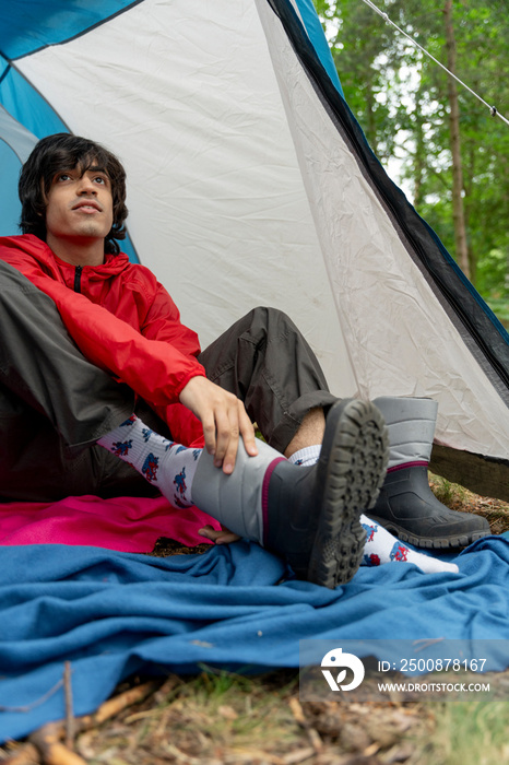 Young man sitting in tent and putting on rain boots