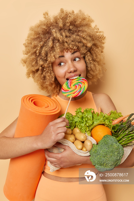 Vertical shot of pensive curly haired woman breaks diet bites lollipop carries fresh vegetables and fitness mat tries to be fit and healthy dressed in activewear isolated over brown background