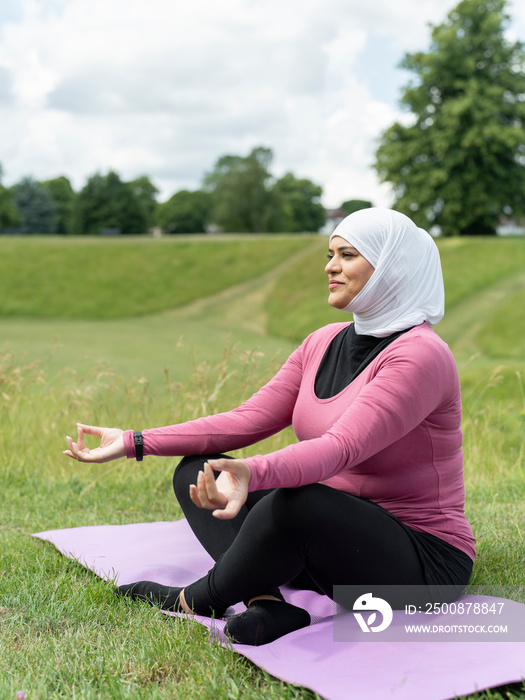 UK,Sutton,Woman in headscarf meditating on yoga mat in grassy field