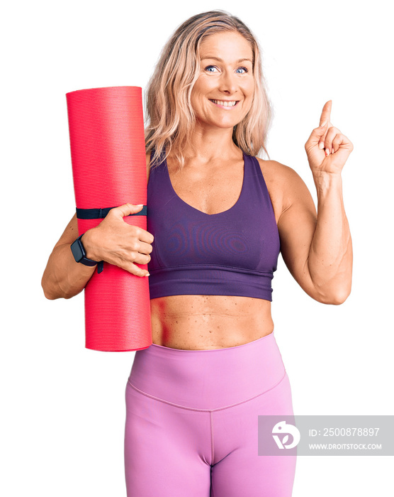 Middle age fit blonde woman holding yoga mat surprised with an idea or question pointing finger with happy face, number one