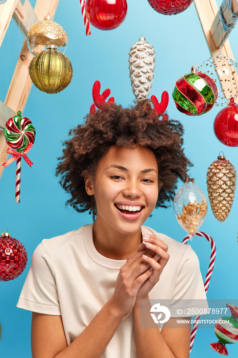 Happy winter holidays and New Year buzz concept. Cheerful young curly woman looks with toothy smile on face poses against blue background with baubles around wears t shirt enjoys decorating room.