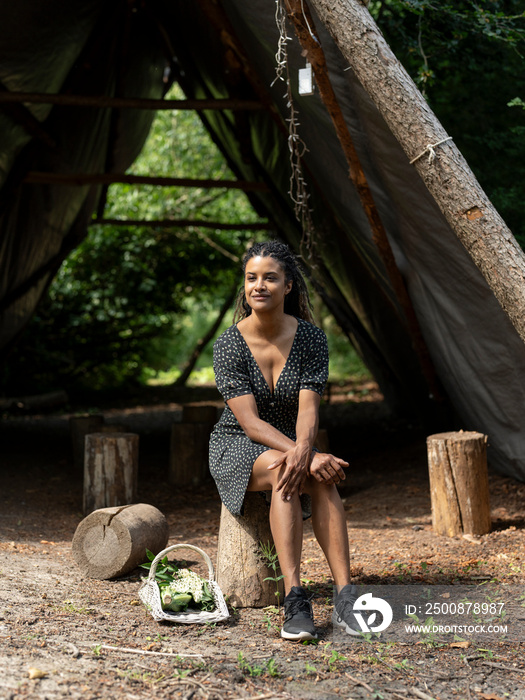 Woman resting on tree stump in front of hut in forest