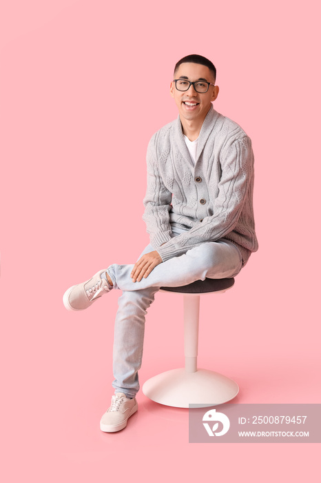 Handsome young Asian man in knitted sweater sitting on chair against pink background