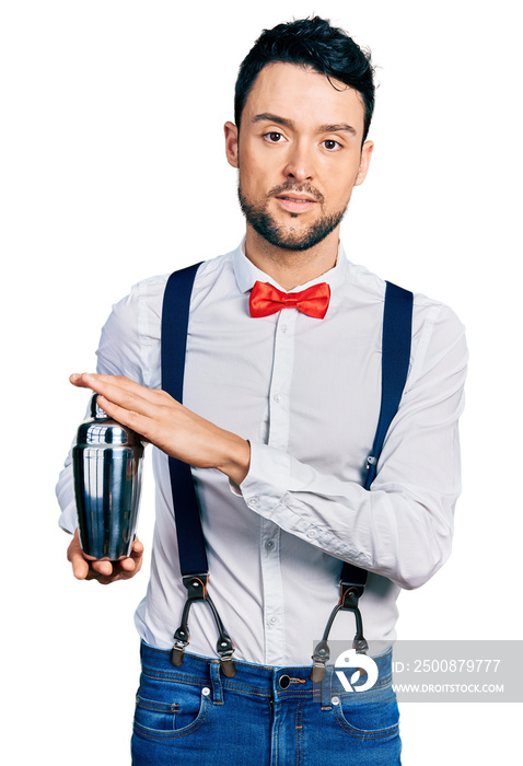 Hispanic man with beard preparing cocktail mixing drink with shaker relaxed with serious expression on face. simple and natural looking at the camera.