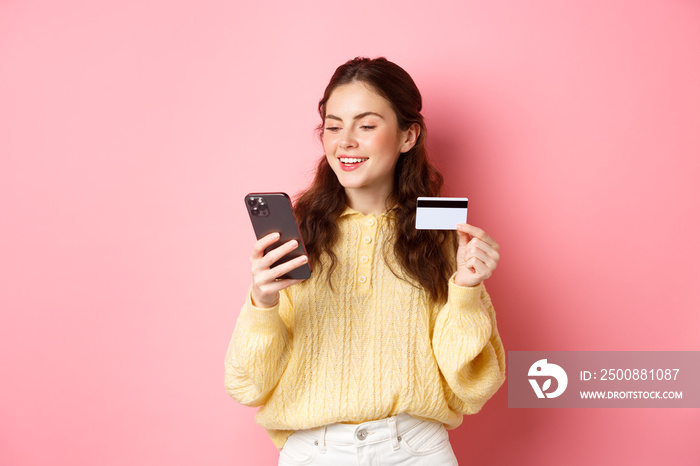 Technology and online shopping. Young pretty lady paying online with credit card, looking at smartphone and smiling, standing over pink background