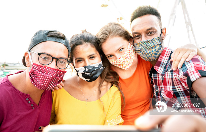 Multicultural milenial travelers taking selfie with closed face masks - New normal travel concept with young people having safe fun together at ferris wheel - Bright warm sunshine filter