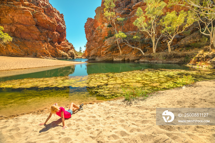 Tourist woman sunbathes on golden sand of Ellery Creek Big Hole at popular waterhole in a gorge surrounded by red cliffs in West MacDonnell Ranges. Tourism in Northern Territory, Central Australia.