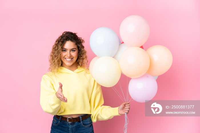 Young blonde woman with curly hair catching many balloons isolated on pink background handshaking after good deal