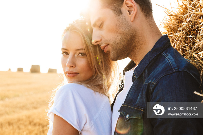 Photo of gorgeous couple man and woman walking on golden field after harvesting, and standing near big haystack