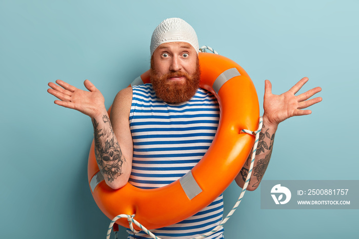 Puzzled bearded man spreads hands with doubt, wears white swimhat, carries lifebuoy, hesitates whether swim on long distance, wears sailor t shirt, stands against blue background. Vacation concept