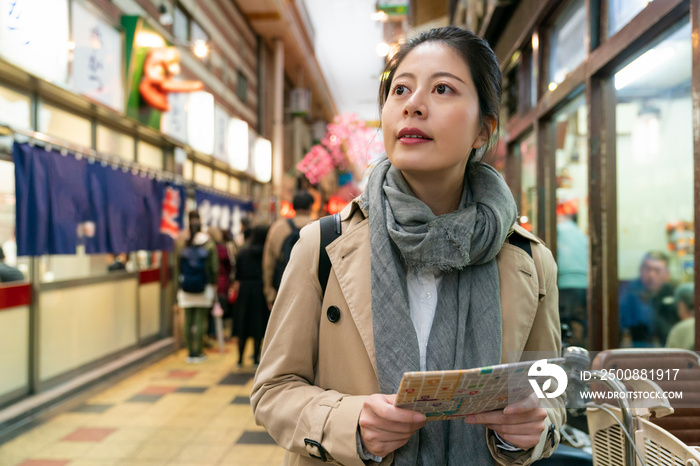 curious Asian Japanese female traveler looking into distance while using guide map to search for restaurant in the arcade food street in Shinsekai area of Osaka Japan
