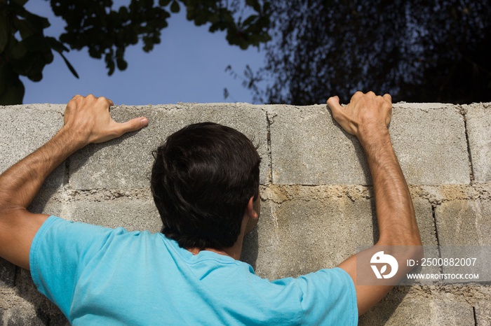 Young man on blue shirt struggling to climb over a grey big brick wall. Illegal immigration, trespassing concept