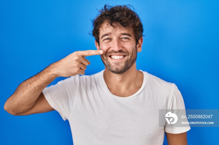 Hispanic young man standing over blue background pointing with hand finger to face and nose, smiling cheerful. beauty concept