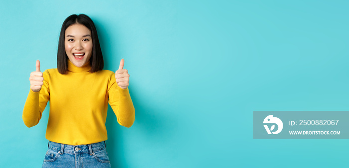 Beautiful asian woman praise good work, showing thumbs up gesture and smiling in approval, recommend product, standing satisfied over blue background