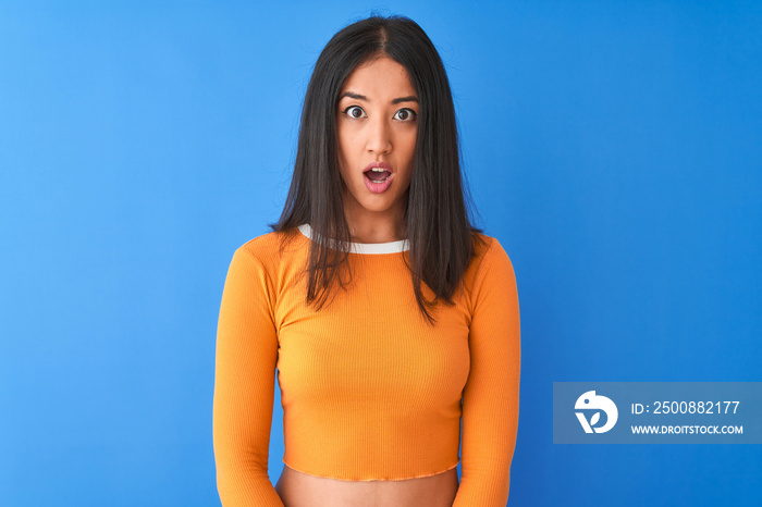 Young beautiful chinese woman wearing orange t-shirt standing over isolated blue background afraid and shocked with surprise expression, fear and excited face.