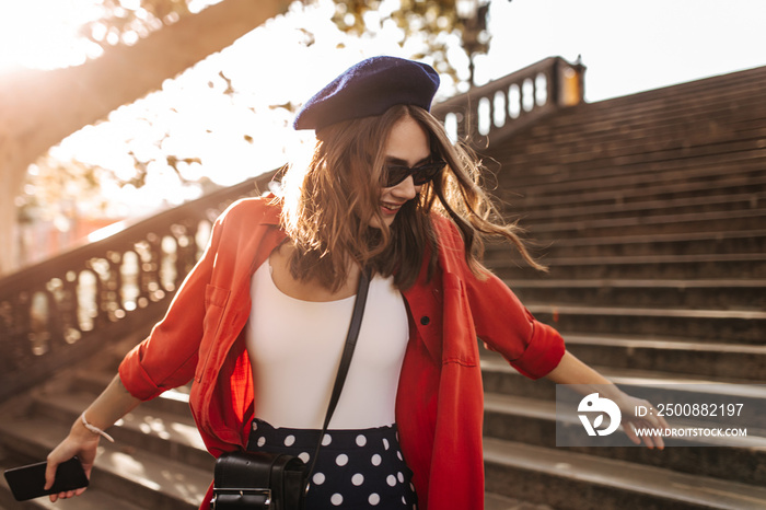 Cute young lady with medium brunette hair, blue beret, stylish sunglasses, white top and red shirt, smiling, posing in sunny autumn city and looking down